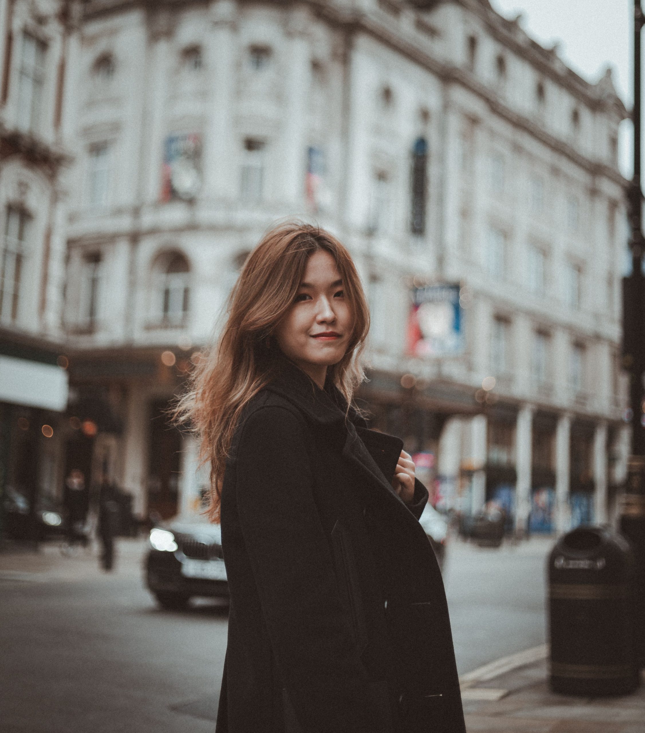 A young woman stands smiling on a London street, wearing a black coat with a historic building in the background.