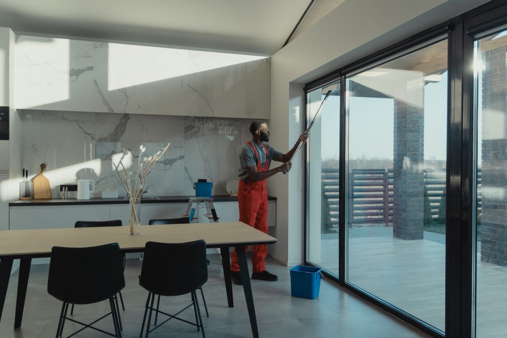 A professional cleaner in uniform washing large glass windows indoors with natural light streaming in.