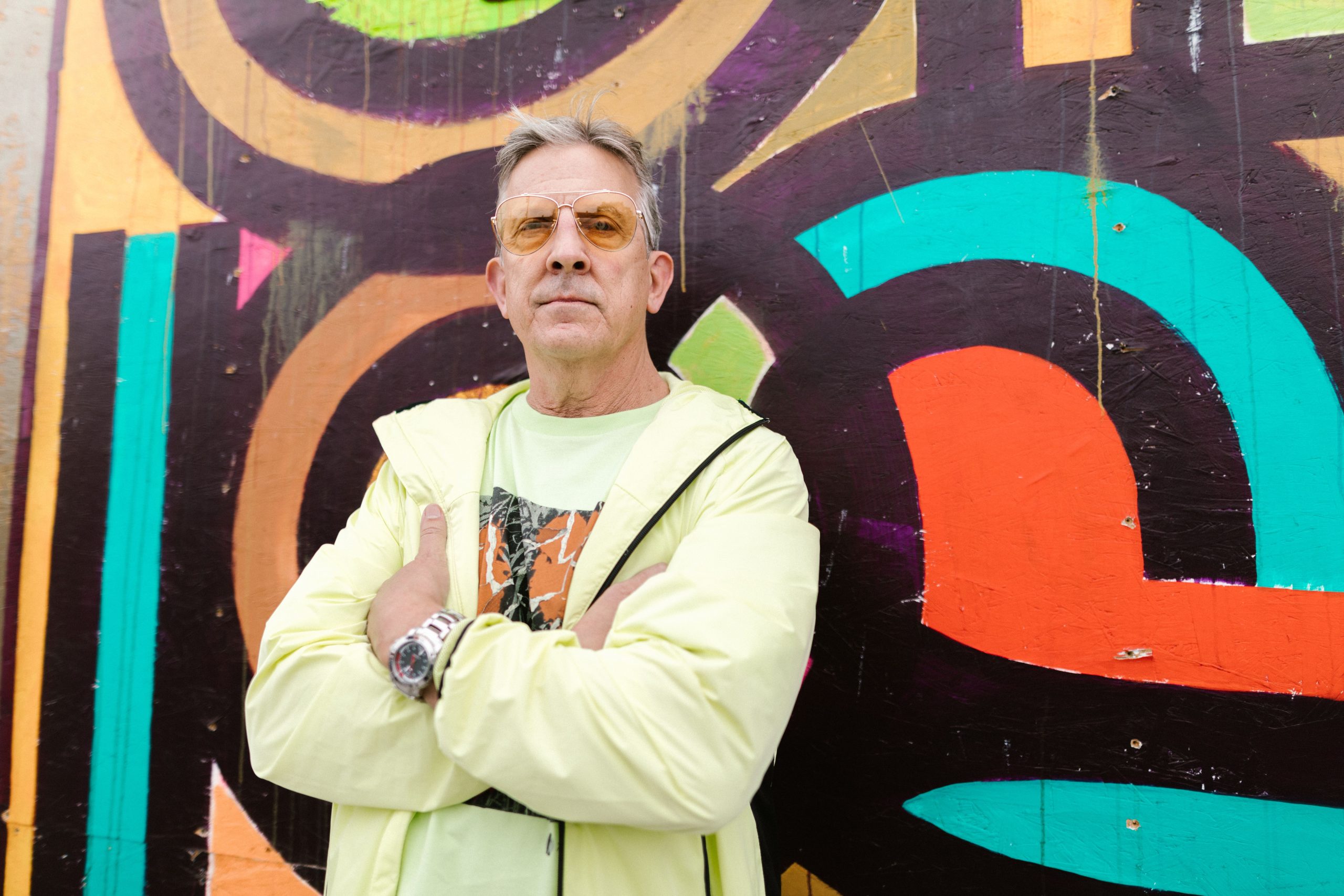 A confident senior man in bright attire poses against vibrant street art, exuding style.
